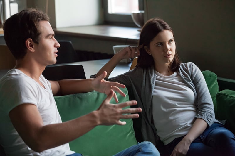 Ein Mann spricht mit einer genervten Frau, während beide auf der Couch sitzen