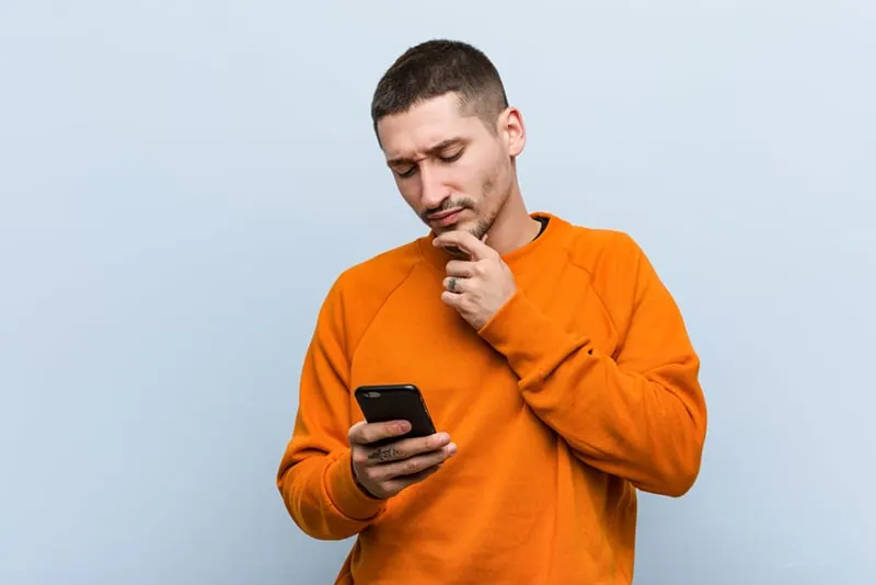 ein Mann, der ein orangefarbenes Sweatshirt trägt, ein Telefon hält und es mit einem zweifelhaften und skeptischen Ausdruck betrachtet