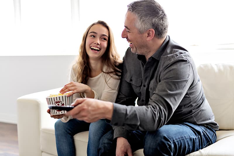 Ein Vater und eine Tochter lachen, während sie auf der Couch sitzen und zusammen fernsehen