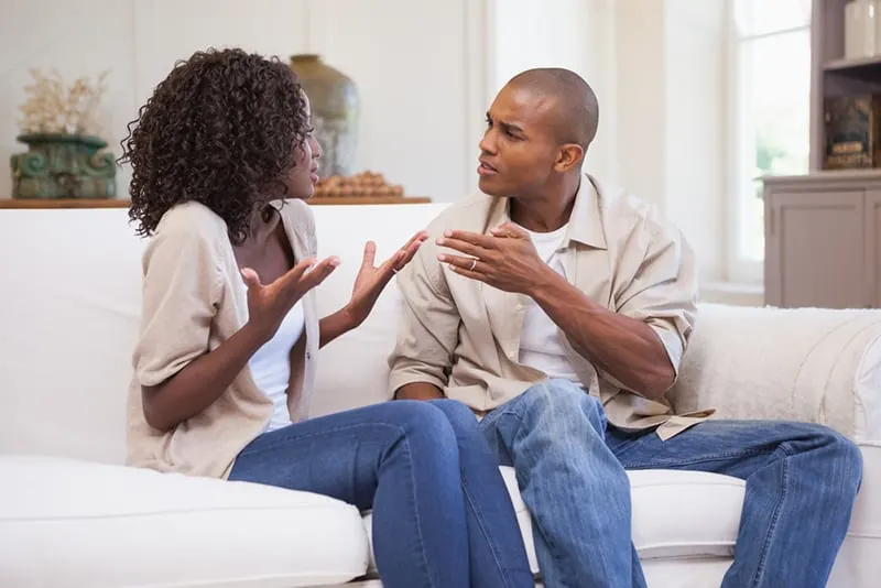 Ein verärgerter Ehemann spricht mit einer Frau, die verwirrt aussieht, während sie auf der Couch sitzt