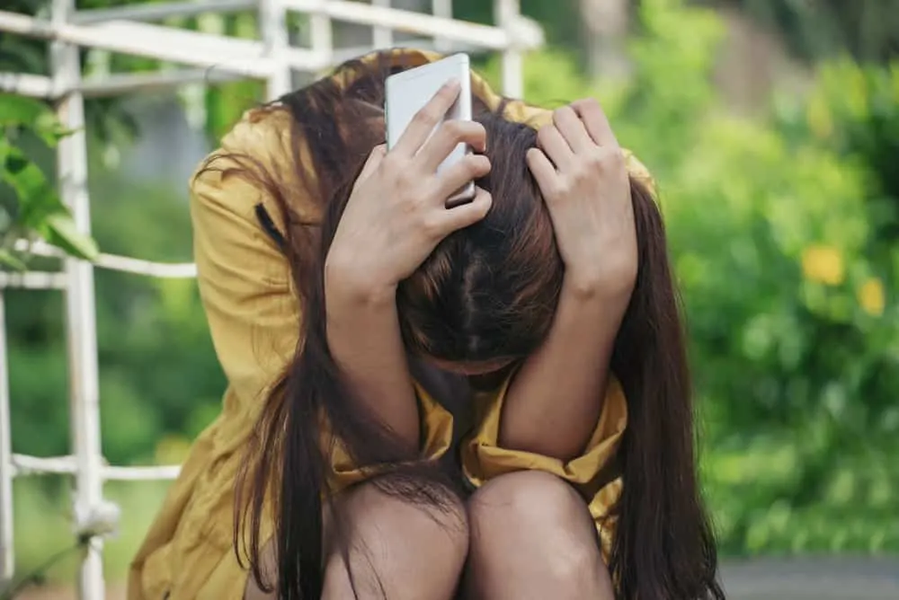 Eine depressive Frau mit einem Handy in der Hand sitzt zusammengerollt auf der Treppe