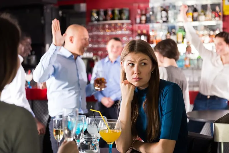 Eine gelangweilte Frau, die am Tisch unter tanzenden Kollegen auf einer Firmenfeier sitzt
