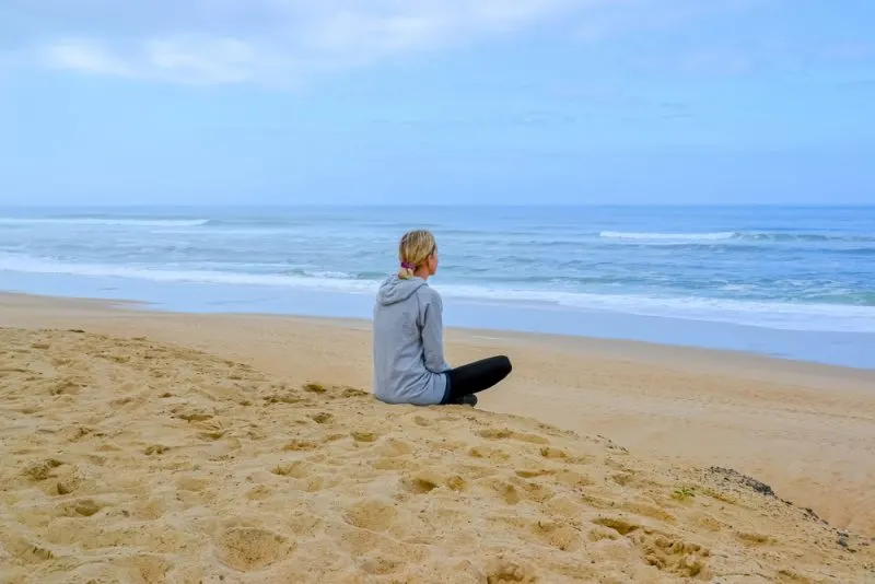 Eine Blondine in einem grauen Sweatshirt sitzt an einem Sandstrand und beobachtet das Meer
