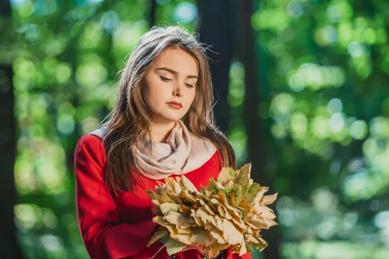 Im Wald steht mit trockenen Blättern in den Händen ein trauriges Mädchen in einem roten Mantel