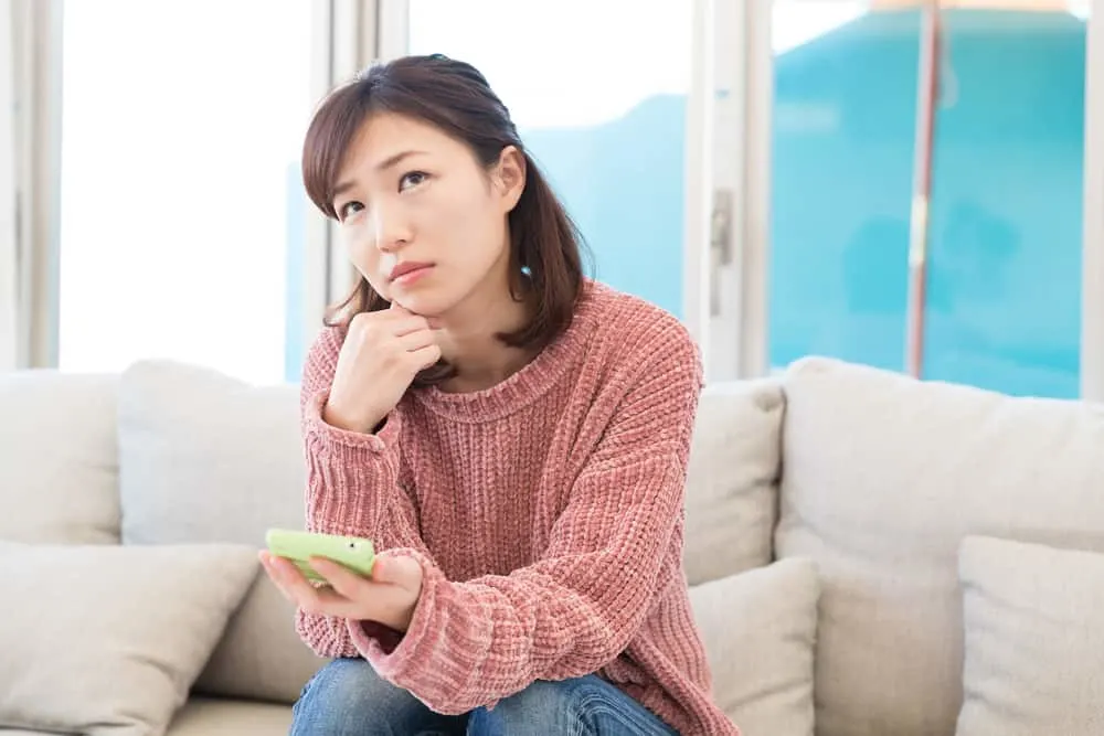 Im Haus auf dem Sofa sitzt eine nachdenkliche traurige Frau mit einem Smartphone in der Hand