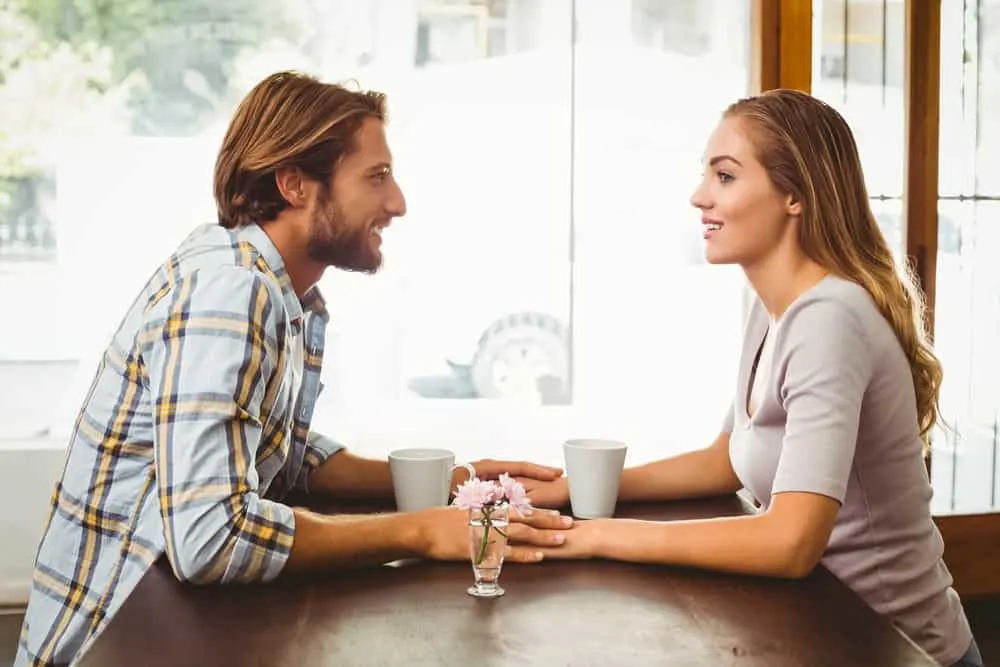 In einem Café an einem Tisch am Fenster sitzen ein lächelnder Mann und eine lächelnde Frau und halten sich an den Händen