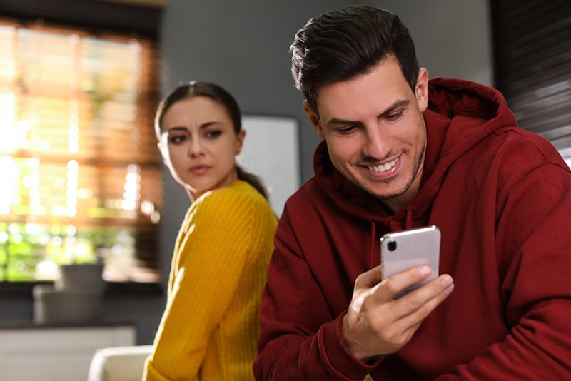 Misstrauische Frau späht in das Smartphone ihres Freundes, während er auf der Couch benutzt
