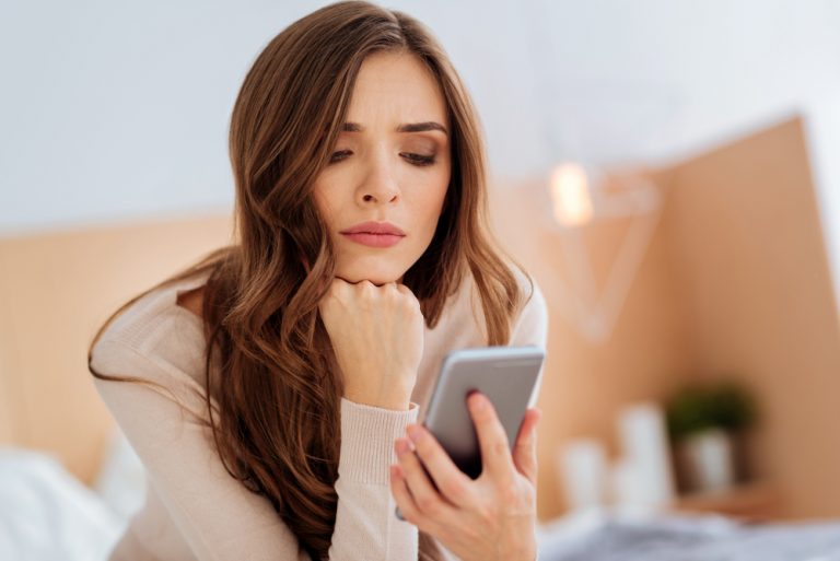 Auf dem Bett im Zimmer sitzt eine besorgte Brünette und benutzt ein Handy