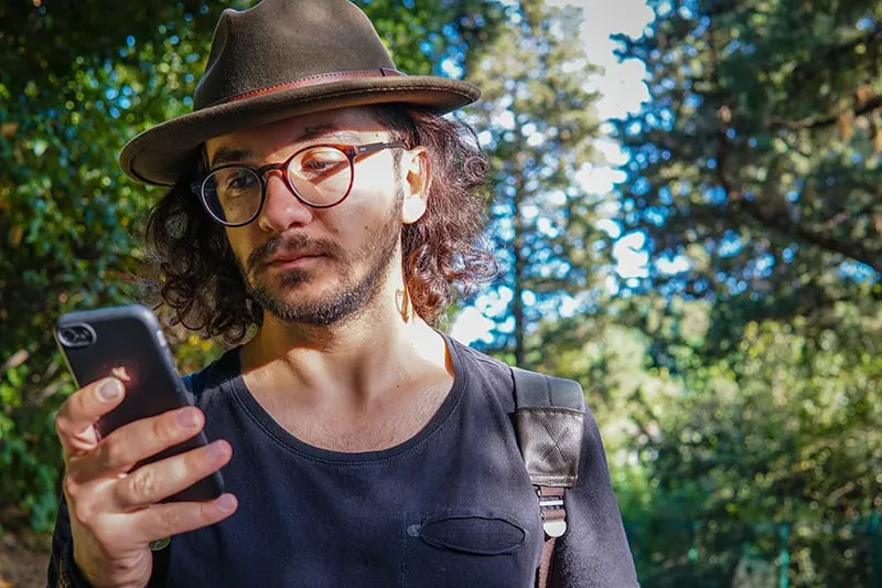ein Mann, der einen Hut trägt, der ein Smartphone beim Gehen im Park betrachtet