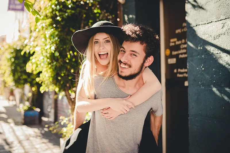 Ein glücklicher Mann, der seine lächelnde Freundin auf dem Rücken trägt, während er auf dem Bürgersteig geht