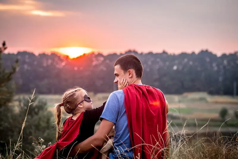 ein kleines Mädchen, das das Gesicht ihres Vaters berührt, während es zusammen auf dem Feld sitzt und beide ein Superheldenkostüm tragen
