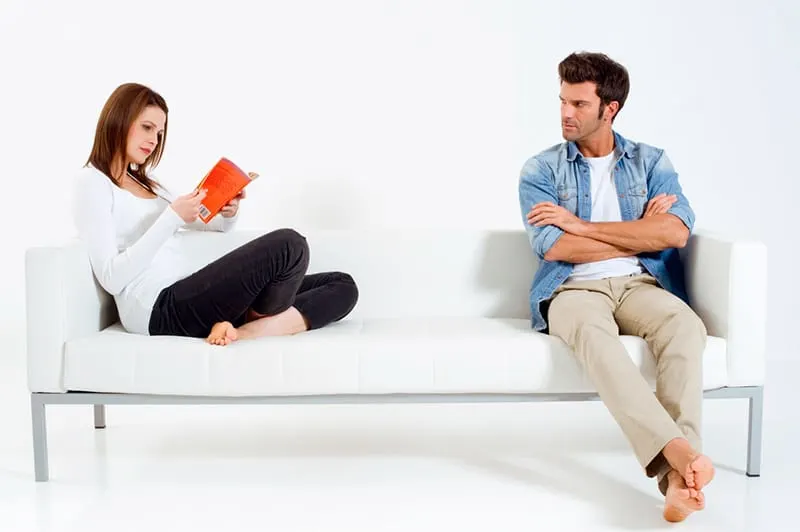 eine Frau, die ein Buch liest, und ihr verärgerter Freund, der sie ansieht, während er getrennt auf der Couch sitzt