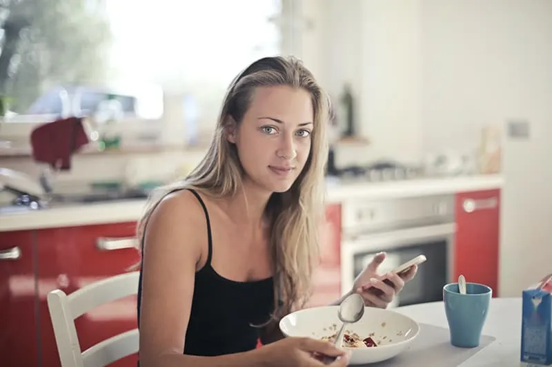 eine Frau, die Frühstück in der Küche isst und Smartphone hält