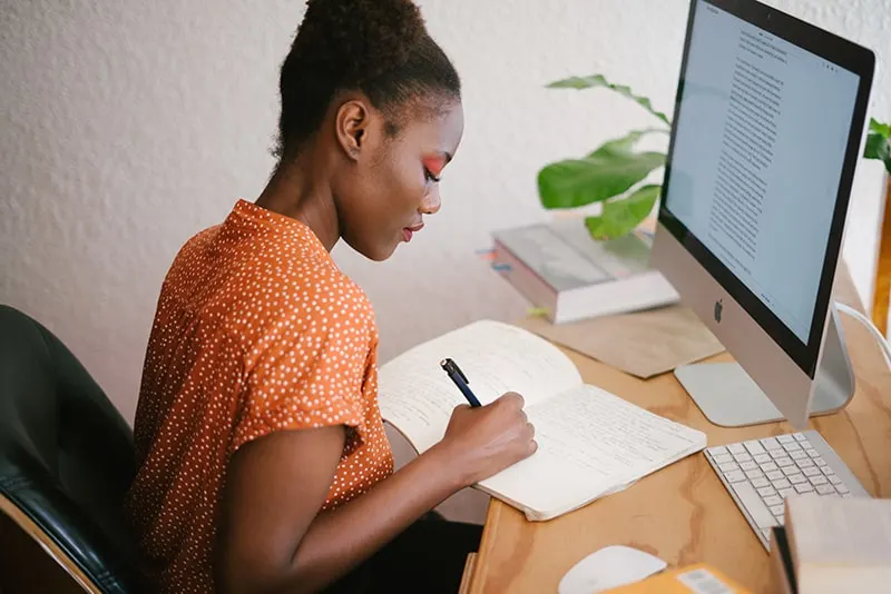 eine Frau, die in ein Notizbuch schreibt, während sie im Büro vor einem Computer sitzt