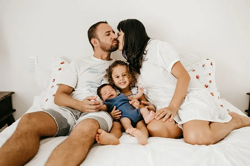 eine Frau küsst ihren Mann auf die Wange, während ihre Kinder zwischen ihnen auf dem Bett sitzen