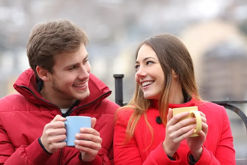 eine lächelnde Frau, die zu einem lächelnden Mann schaut, der ihr Kompliment macht, während sie zusammen Tee im Freien trinkt