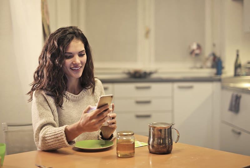 eine lächelnde Frau, die ein Smartphone benutzt, während sie in der Küche sitzt