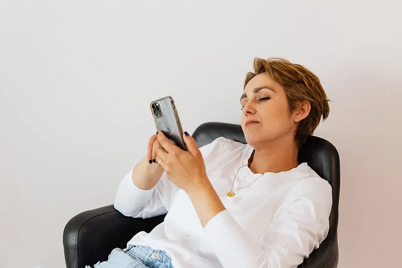 eine nachdenkliche Frau, die ein Smartphone betrachtet, während sie im Sessel sitzt