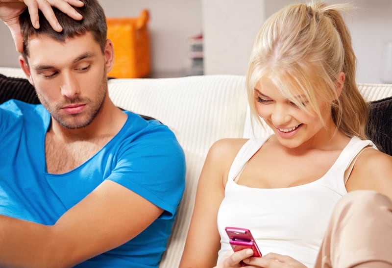 lächelnde Frau mit ihrem Smartphone und ihrem Freund, der sie überprüft, während er auf der Couch sitzt