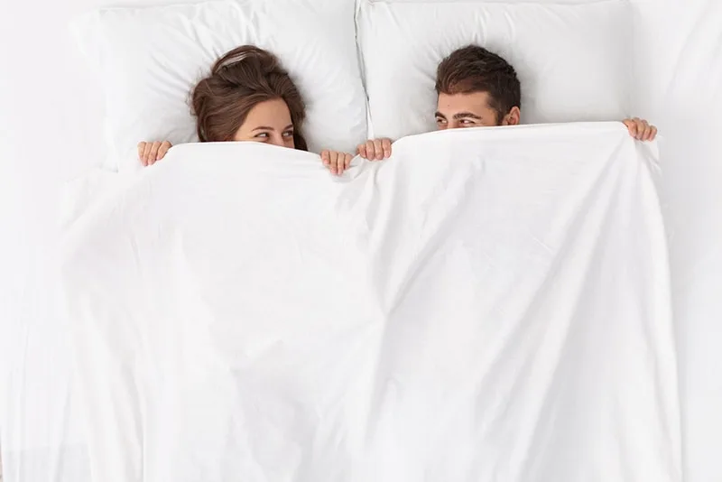 lächelndes Paar, das mit weißen Laken bedeckt, während es in einem Bett liegt