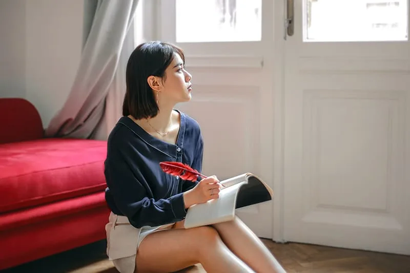 nachdenkliche Frau, die in einem Notizbuch schreibt, während sie zu Hause auf dem Boden sitzt