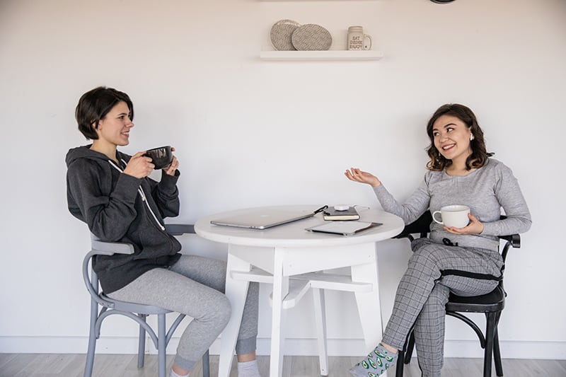 zwei lächelnde Freundinnen, die sprechen, während sie zusammen Kaffee am Tisch trinken