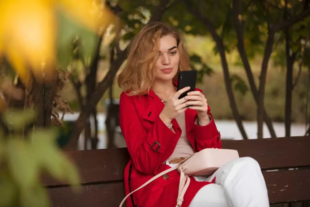 Auf einer Holzbank im Park sitzt ein Pavuša in einem roten Mantel und schreibt eine SMS