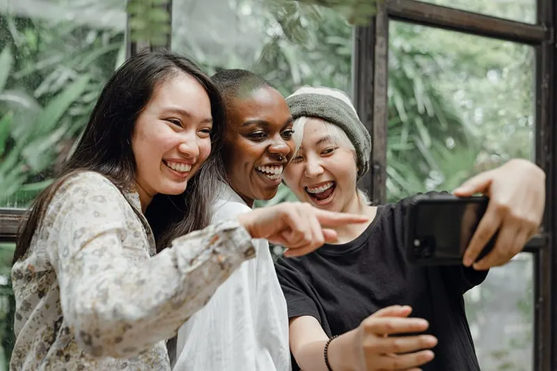 Drei lächelnde Frauen machen ein Selfie, während sie in der Nähe des Fensters stehen