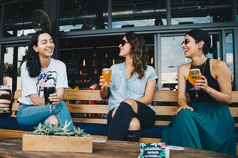 Drei lächelnde Frauen sitzen auf der Bank und halten Getränke in der Kneipe