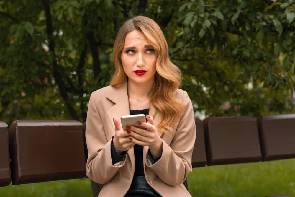 Im Park sitzt eine Blondine mit rotem Lippenstift und benutzt ein Smartphone