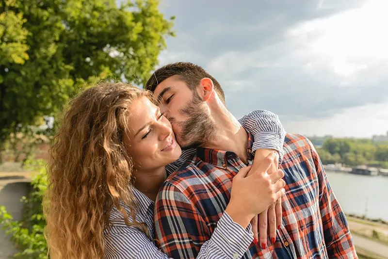 Mann küsst eine Frau auf eine Wange, während sie ihn von hinten umarmt
