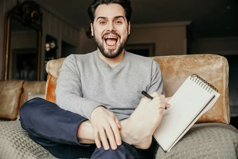 Mann schreibt einen Text in das Notizbuch mit den Zehen des Fußes, während er auf der Couch sitzt