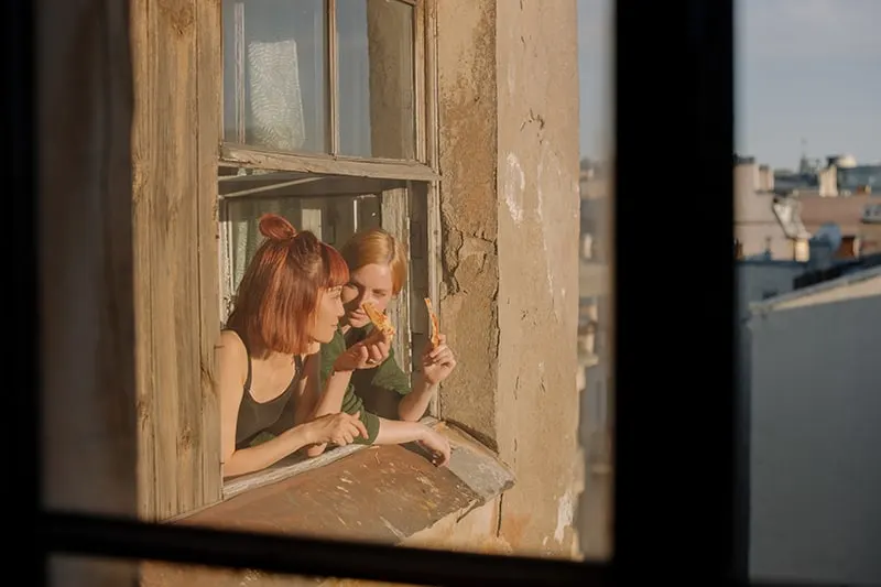 Zwei Frauen lehnen sich an das Fenster eines alten Gebäudes und essen Pizza