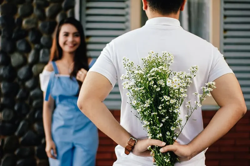 ein Mann, der einen Blumenstrauß hinter seinem Rücken hält, während er vor lächelnder Frau steht
