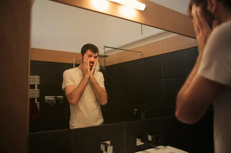 Ein Mann, der sich selbst in den Spiegel schaut und dabei das Gesicht mit den Händen berührt