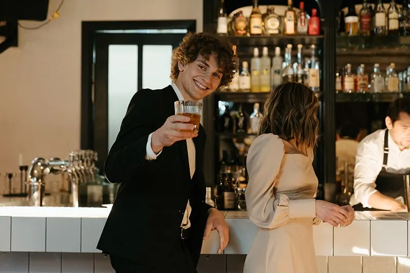 ein lächelnder Mann, der einen Drink hält, während er neben einer Frau steht, die sich an die Theke lehnt