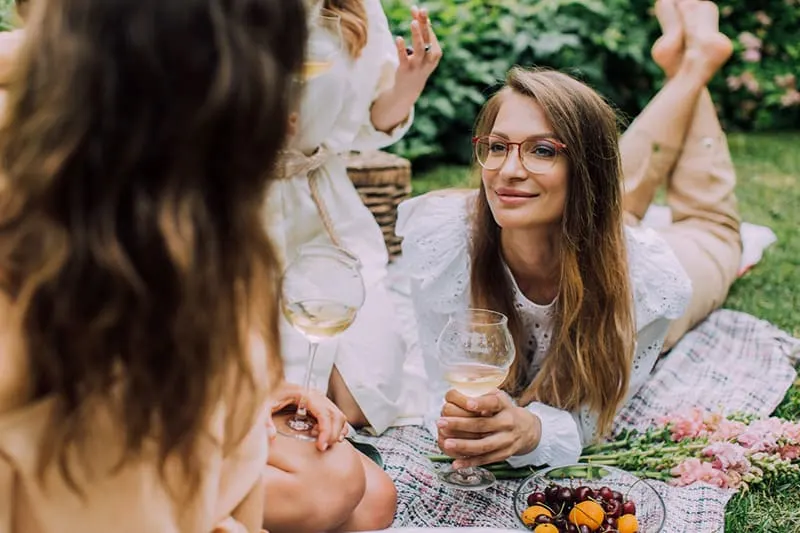 eine Frau, die auf einer Decke liegt und ihre Freundin ansieht, während sie zusammen Wein trinkt