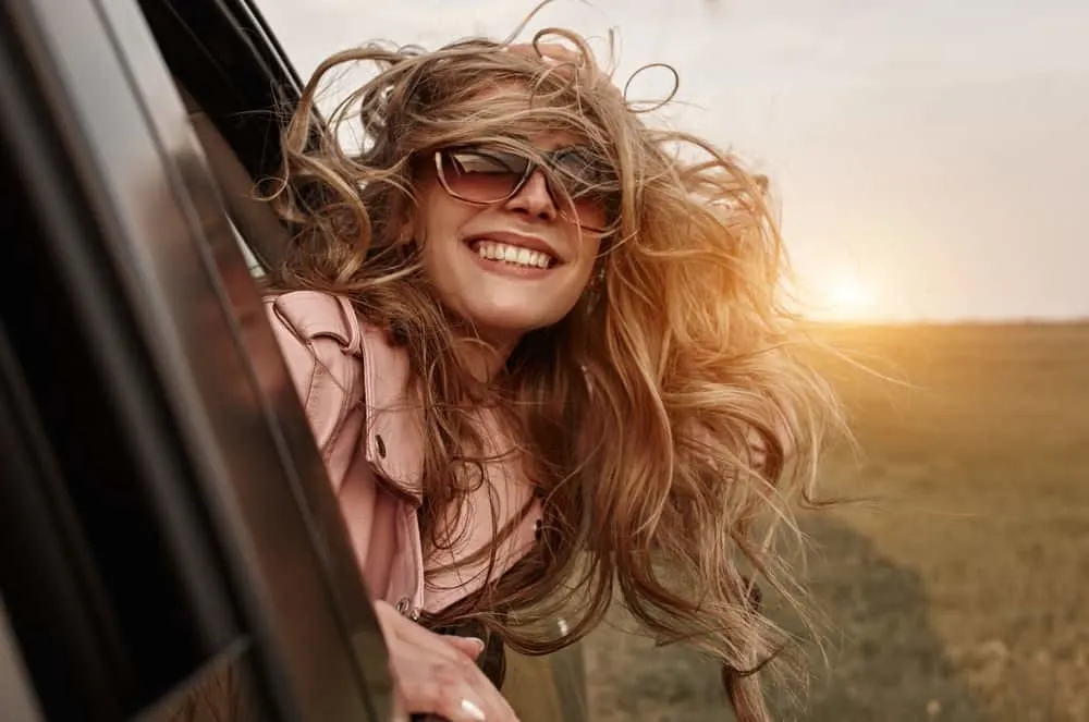 eine lächelnde Blondine mit Brille, die sich während der Fahrt aus einem Autofenster lehnt