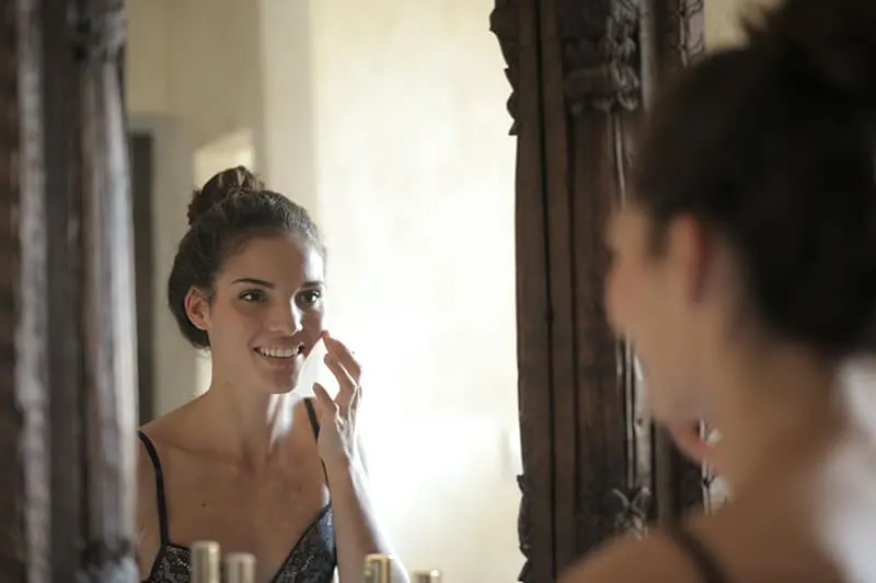 eine lächelnde Frau, die Creme auf Gesicht vor dem Spiegel aufträgt