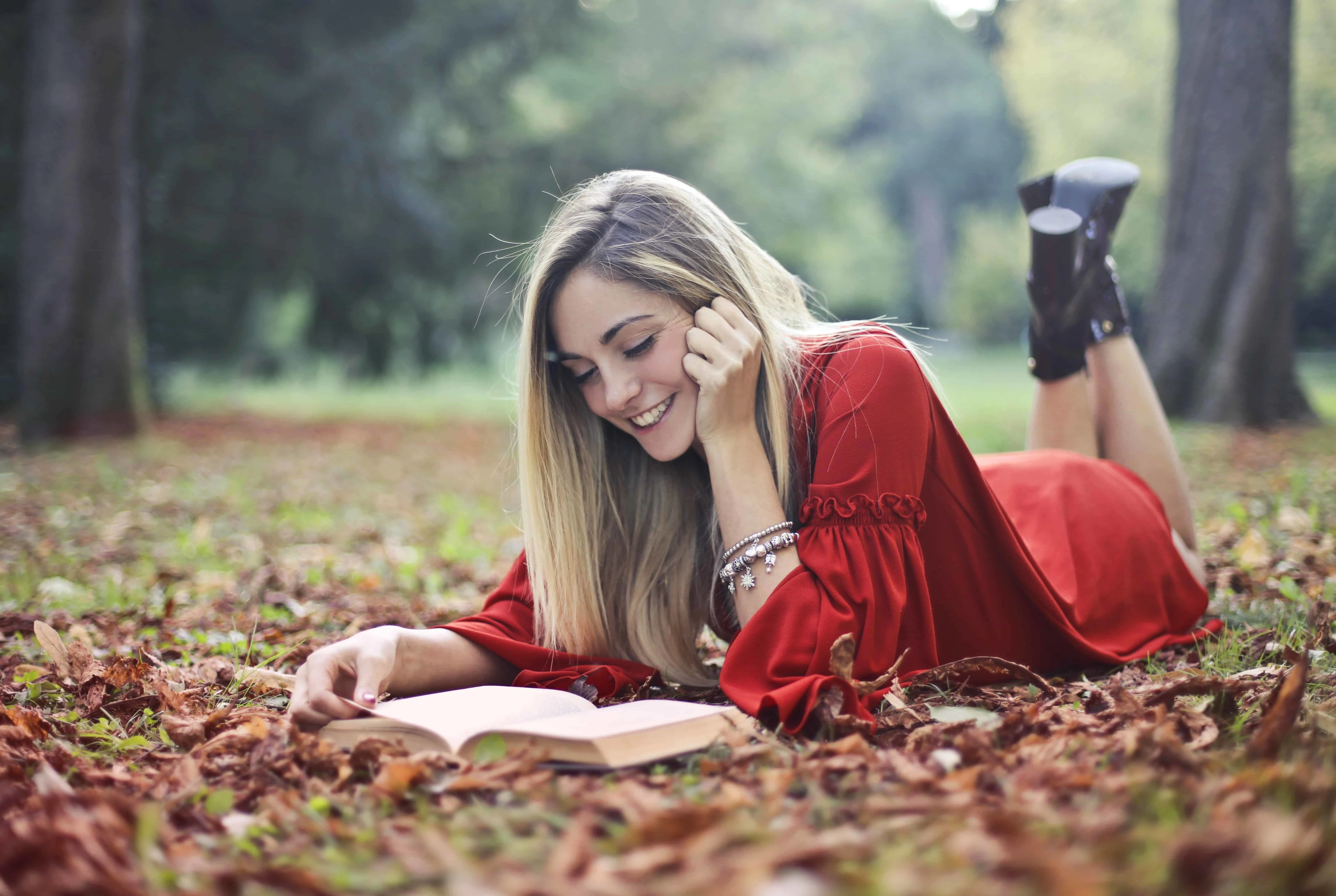 eine lächelnde Frau, die ein Buch liest, während sie auf getrockneten Blättern auf dem Boden liegt