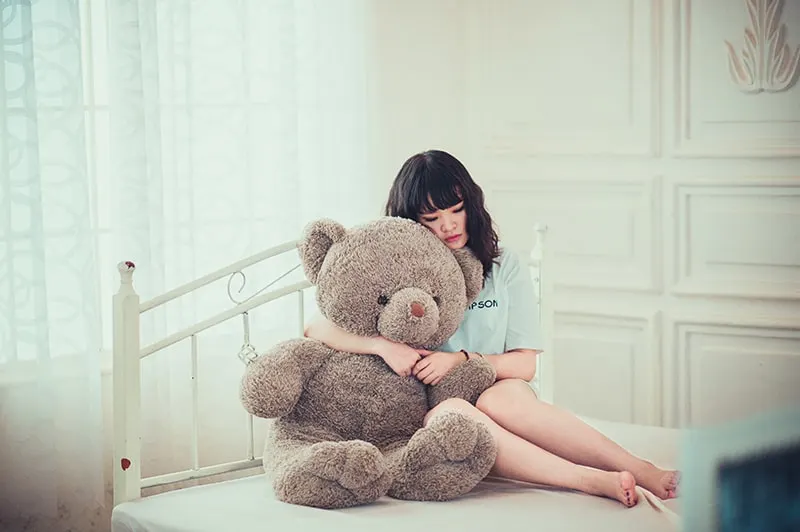 eine traurige Frau, die einen Teddybären umarmt, der auf dem Bett sitzt