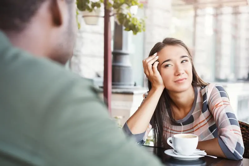 gelangweilte Frau, die einen Mann betrachtet, der über ihr sitzt, während Kaffee zusammen auf einem Date trinkt