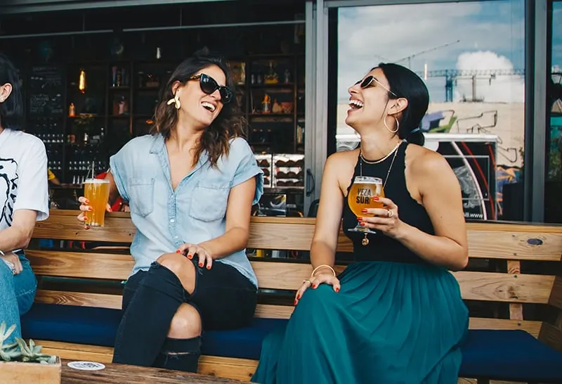 zwei lachende Frauen, die Getränke halten, während sie auf der Holzbank sitzen