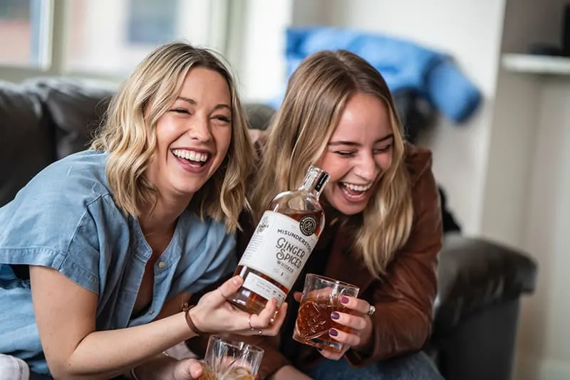 zwei lachende Frauen, die Whisky trinken, während sie auf der Couch sitzen