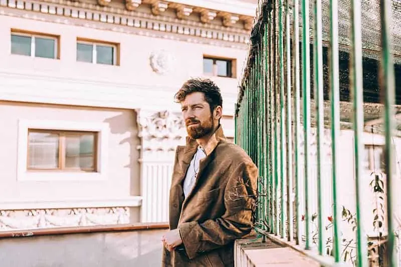 Ein Mann mit Bart steht in einem braunen Mantel am Zaun