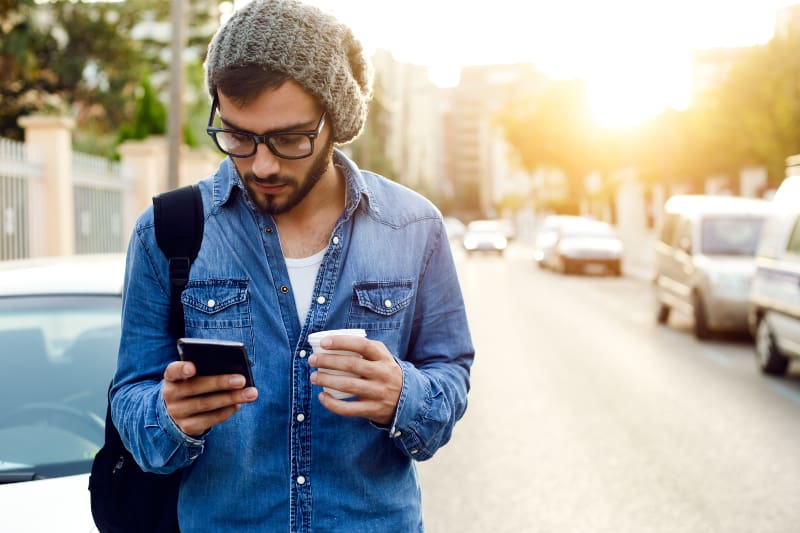 Ein junger Mann hält einen Kaffee in der Hand und tippt eine Nachricht auf sein Handy