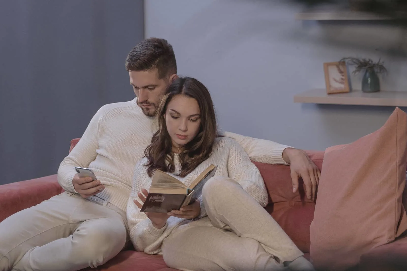 Eine Frau liest ein Buch und ein Mann benutzt sein Smartphone, während sie zusammen auf der Couch sitzen