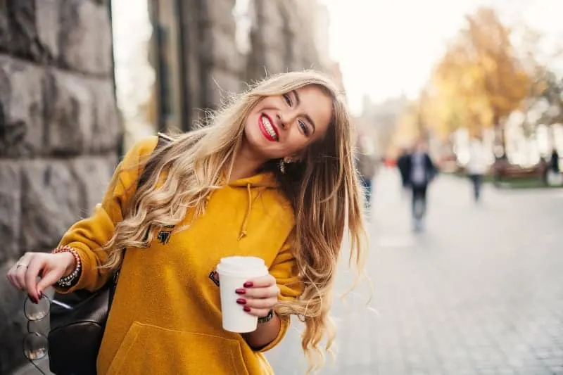 Eine schöne Blondine in einem gelben Sweatshirt lacht und hält einen Kaffee in der Hand