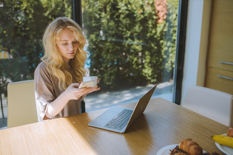 Frau hält eine Tasse Kaffee, während sie ihr Smartphone benutzt und vor einem Laptop sitzt