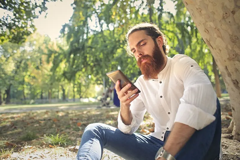 Ein Mann, der auf einem Smartphone tippt, während er alleine im Gras im Park sitzt